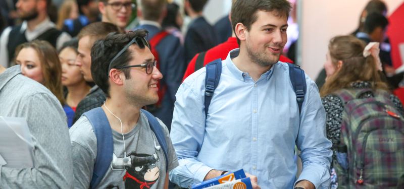 Zwei junge Männer mit Rucksäcken gehen lächelnd durch einen vollen Messegang