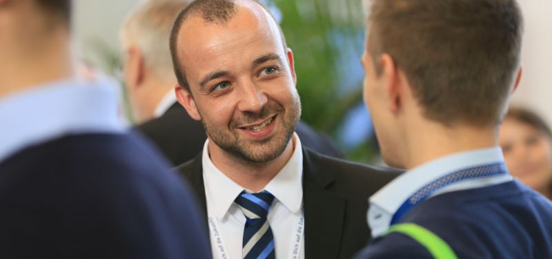 Ein lächelnder Aussteller im Anzug hört einem jungen Besucher aufmerksam zu